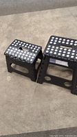Photo of two black folding step stools of different sizes placed on a concrete floor, showing the top non-slip dotted surface and side leg structure.