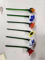 Top view of six hand blown glass flowers laid out on a white surface showing different flower shapes and colors including orange, blue and white, red, yellow, and red bell shapes.