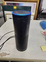 Full view of Amazon Echo smart speaker standing on a white surface with its blue light ring on, showing some scratches and scuffs.