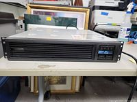 Front view of black rack-mountable UPS unit with ventilation grille and small display showing output status.