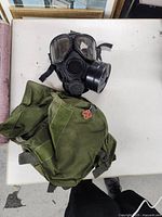 Black military gas mask with attached round filter canister and clear eye lenses alongside the green carrying bag placed on a white table.
