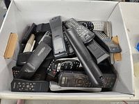 Photo showing a white box filled with multiple used remote controls in various sizes and brands, mainly black and gray in color.