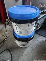 Two stacked 3.5 gallon buckets with blue lids and white labels in a garage environment.