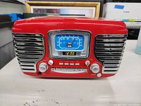 Front view of Crosley red CD player and radio with chrome speaker grills, analog-style tuning dial with blue face, digital clock display, two large knobs, and buttons under the dial.