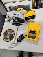 DeWalt cordless circular saw with battery compartment, charger base, power cord, circular saw blades, and metal guide attachment on white table.