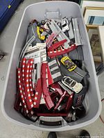 Bin filled with red and black slot car track pieces with checkered guardrails and three slot cars visible on top.