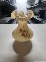 Front view of the cream-colored glass vase showing the hand painted floral design on the rounded body and the ruffled rim at the top.