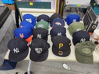 Front view of hats arranged on a table showing logos of New York Yankees, Brooklyn Nets, New York Jets, Denver Broncos, Pittsburgh Pirates, New York Mets.