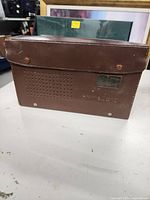 Front view of brown leather carrying case with perforated speaker holes and metal nameplate with embossed markings.