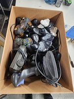 Box of assorted wired and wireless game controllers and joysticks including Xbox 360 controllers and flight joystick throttle stick.