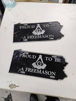 Top side of two black Freemason vanity plates with white text and emblem.