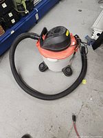 Side and front angle shot of the Rigid shop vac showing the 9-gallon white drum, orange lid, black motor housing, and attached black hose.