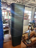 Vertical stack of 28 black plastic rectangular trash cans with rounded corners and horizontal ridges, placed indoors among various household items.