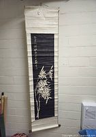Full length photo showing the hanging scroll mounted on a wall. Scroll features a black ink wash panel with white bamboo design and calligraphy, surrounded by light colored fabric with visible folds and minor wear.