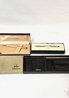 Three boxed ballpoint pens and one boxed black leather wallet displayed on white background.