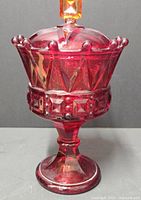 Front view of ruby red glass footed candy dish with lid showing detailed faceted design, crown rim, and square finial on lid.