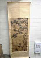 Full view of the hanging scroll showing the traditional landscape painting mounted on beige textile backing, hanging against a white brick wall.