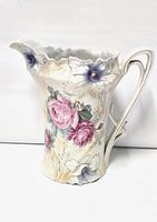 Front and side views showing pink and purple floral hand-painted designs on a white satin finish porcelain milk pitcher with scalloped rim and ornate handle.