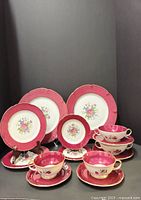 Full view of all dinnerware pieces in the set: 2 dinner plates, 2 side plates, 2 handled soup bowls with plates, 2 dessert plates, 2 cups and saucers. Displayed on stands showing red borders and floral centers.
