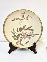 Front view of the round ceramic plate with floral design and gold gilded rim on a dark wooden stand.