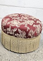 Side angled view of the footstool showing the cherub patterned red fabric and gold bullion fringe trim against a white brick wall and concrete floor.