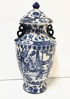 Front angle of the tall porcelain jar showing the detailed blue and white painted scene of people and architecture, dark handles, and removable lid with floral design.