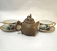 Tea set showing two porcelain cups and saucers with hand-painted traditional decor in front of small decorative clay teapot with attached lid chain