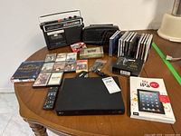 Full lot items laid out on a round wooden table shows the LG Blu-ray player, remote, vintage radios and cassette players, multiple cassette tapes, CDs, DVDs, and an iPad book.