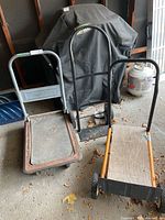 Three items in garage: two metal frame carts including one gray and one orange with wooden base, plus one black-handled hand truck with rubber tires.