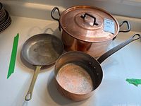 Three copper cooking vessels on a kitchen counter including a large copper pot with lid, a small saucepan without lid, and a fry pan.