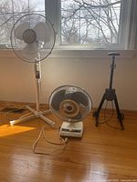 Three items displayed on hardwood floor against window: adjustable height floor fan, smaller desk fan, and luggage cart.