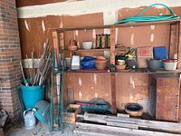 Wide view of wooden shelving with various plastic and ceramic pots, gardening tools, metal trellis frames, wooden dowels, and garden hose in bucket container.