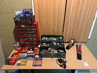 Wide view of tabletop displaying two hand planers, a green tackle box-style toolbox filled with hardware, black Mastercraft glue gun, red and black hardware storage organizers with assorted boxes and hardware items laid out including screws and nails.