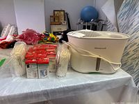 Honeywell humidifier, wrapped filters, and boxes of demineralization cartridges on a table with a background of some household items.