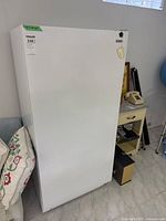 White upright Kenmore freezer showing full front door and adjacent room furniture (pillows and shelf).
