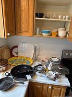 Kitchen counter and shelves showing a variety of dishes, bowls, fry pans including a cast iron pan, serving platters, and wood cutting boards.