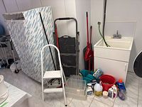 Laundry and cleaning area showing clothes rack with zippered cover, two folding step ladders, mop buckets, cleaning solvent bottles and brushes next to utility sink