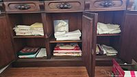 Wooden cabinet with shelves containing folded damask tablecloths, cloth napkins, and placemats.