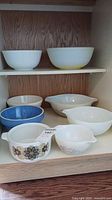 Shelf with two large white and yellowed mixing bowls, one large white bowl shows discoloration, and smaller bowls underneath include floral-patterned Pyrex and ceramic bowls in white, blue, and striped designs.