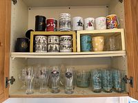 Two shelves of mugs, tumblers and beer glasses inside a wooden cabinet. Top shelf has assorted mugs and tumblers including floral pattern mugs and MCM turquoise tumblers, and an ice bucket. Bottom shelf has several clear glass beer glasses with Upper Canada Brewing Company labels.
