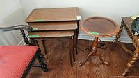 Photo showing the set of three wooden nesting tables stacked together next to a red upholstered chair and the tri-legged round wooden stand.