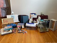 Overall lot of office and art supplies on hardwood floor: hand-cranked shredder, DVDs, paper stacks, briefcase, keyboards, batteries, and watercolor supplies.