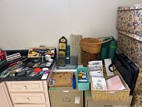 Photo showing full assortment of office supplies and miscellaneous items arranged on a cabinet and floor including baskets, three-hole punch, calculators, keyboard, briefcase, and other items.