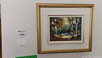 Framed oil painting depicting forest landscape. Shows gold and white frame with signature on painting.