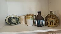 Overview photo showing two artisan pottery vases, a pottery bowl, and two mugs arranged on a white shelf.