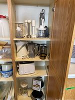 Kitchen cabinet shelf showing all appliances in lot: Kenwood immersion chopper and attachments, Betty Crocker electric kettle, coffee percolator, Toastmaster toaster on middle shelf, Cuisinart waffle maker with manual on bottom shelf.