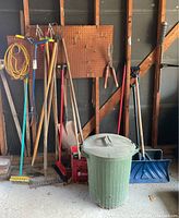 Full view of the yard and garden tools arranged in a garage showing items described including garden hoe, shovels, rake, broom, and trash bin.