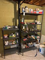 Photo showing two metal shelving units against a basement concrete cinder block wall with various small items on the shelves. The units have a dark finish and show moderate signs of wear.