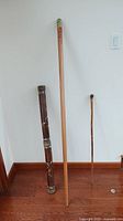Three sticks photographed standing upright against a white wall: a dark brown carved bamboo rain stick on the left, a tall carved wooden walking stick in the center, and a smaller carved bamboo walking stick on the right.