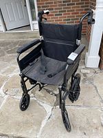 Front angle view of black Airgo Comfort-Plus XC lightweight transport wheelchair showing fabric seat with seatbelt, padded armrests, and large rear wheels.